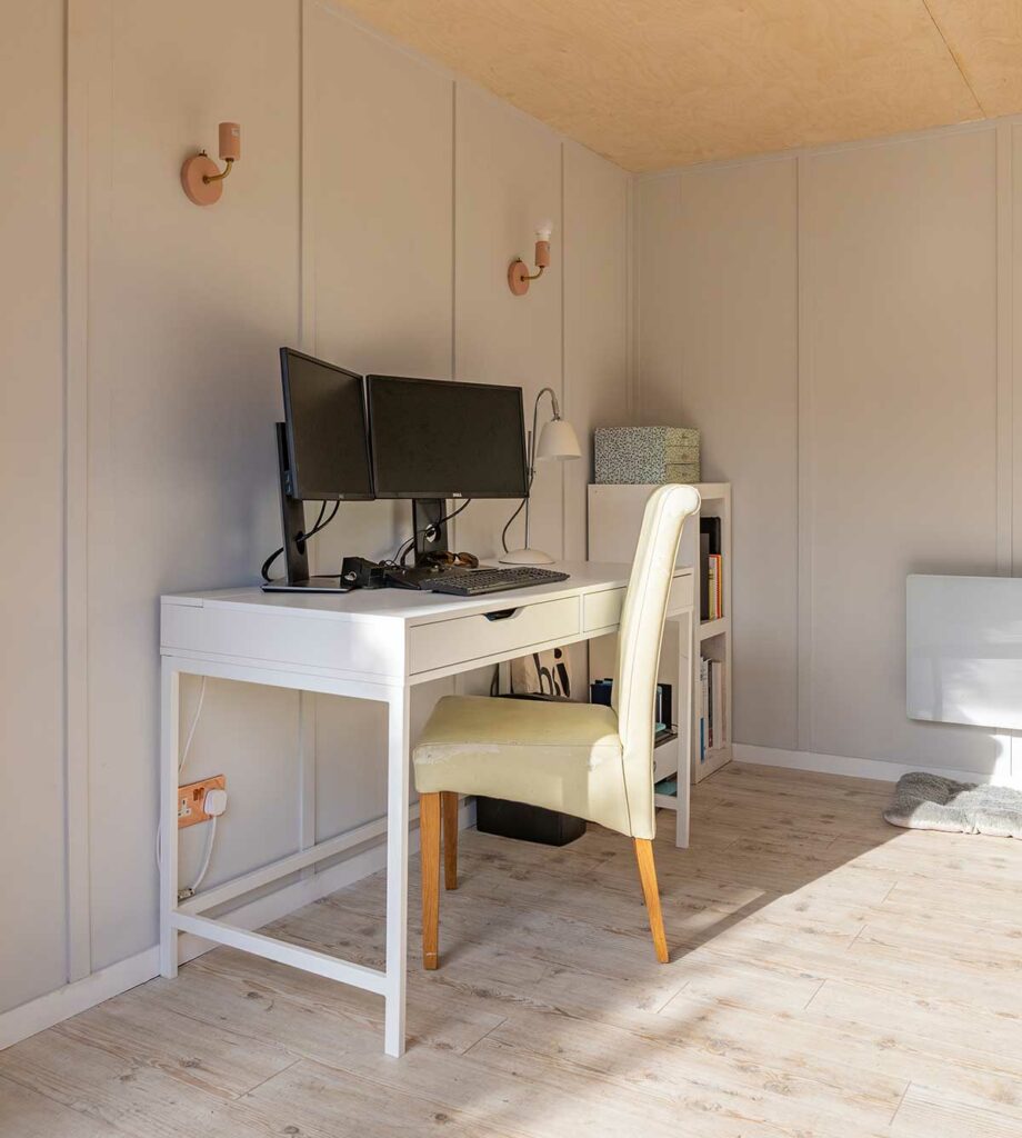 A bright, minimalist home office in the style of modern garden rooms features a white desk with two monitors, keyboard, lamp, and mouse. A light-colored chair sits at the desk, with a small bookshelf and wall lights in the background.