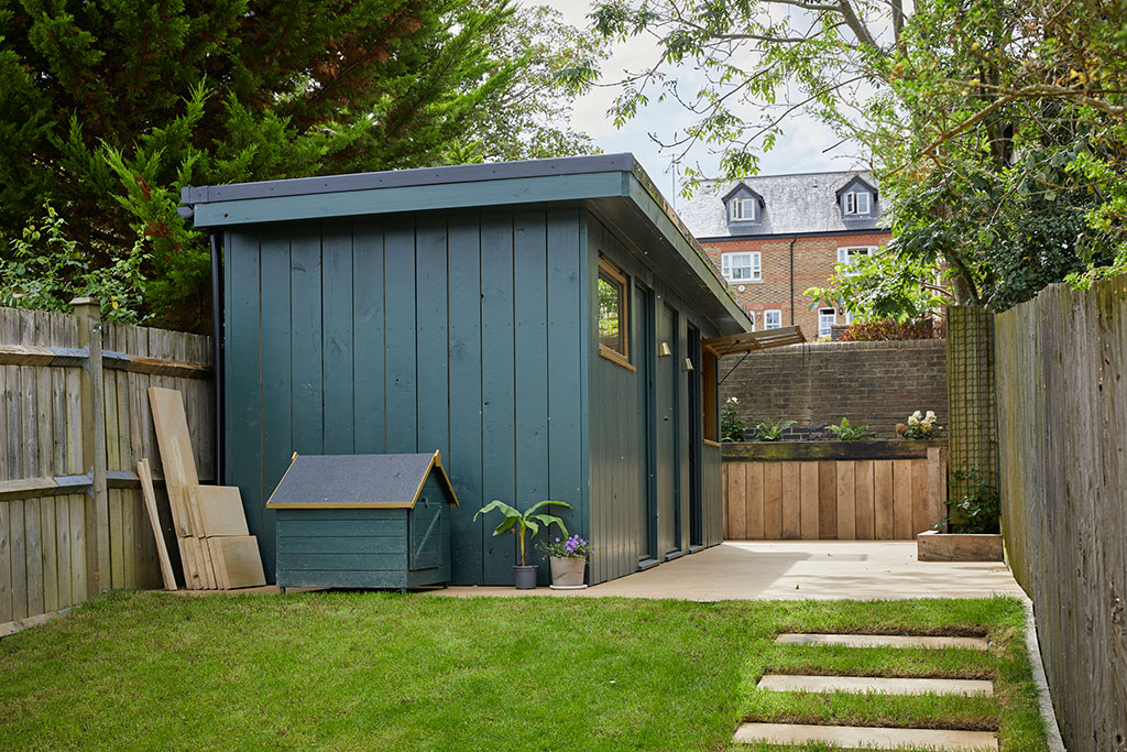 A small, blue wooden shed with a matching doghouse sits in a tidy backyard. The shed is surrounded by a green lawn, potted plants, wooden fencing, and trees, with houses visible in the background.